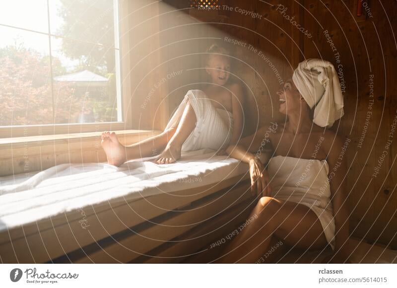 Mutter und Tochter in einer Sauna, die Mutter mit einem Handtuch auf dem Kopf, beide lächelnd und sitzend Kind Familie Wellnessbad Resort Hotel Fenster Schweiß