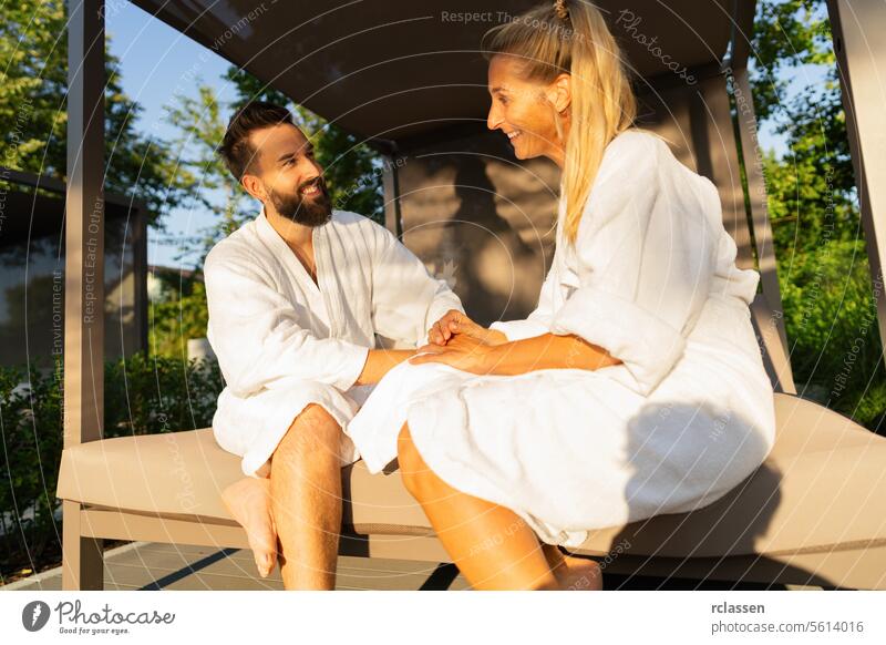Lächelndes verliebtes Paar in weißen Bademänteln auf einer Liege im Freien im Sonnenlicht im Spa-Wellness-Hotel sitzend Bankliege weiße Gewänder Außenbank