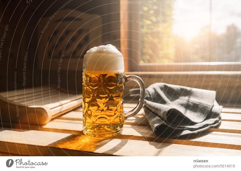 Bierkrug mit schäumendem Bier ruht auf einem Sims in einer Sauna und fängt Sonnenlicht ein. In der Nähe finnische Sauna Hüte auf einer Holzbank. Spa und Wellness-Konzept Bild