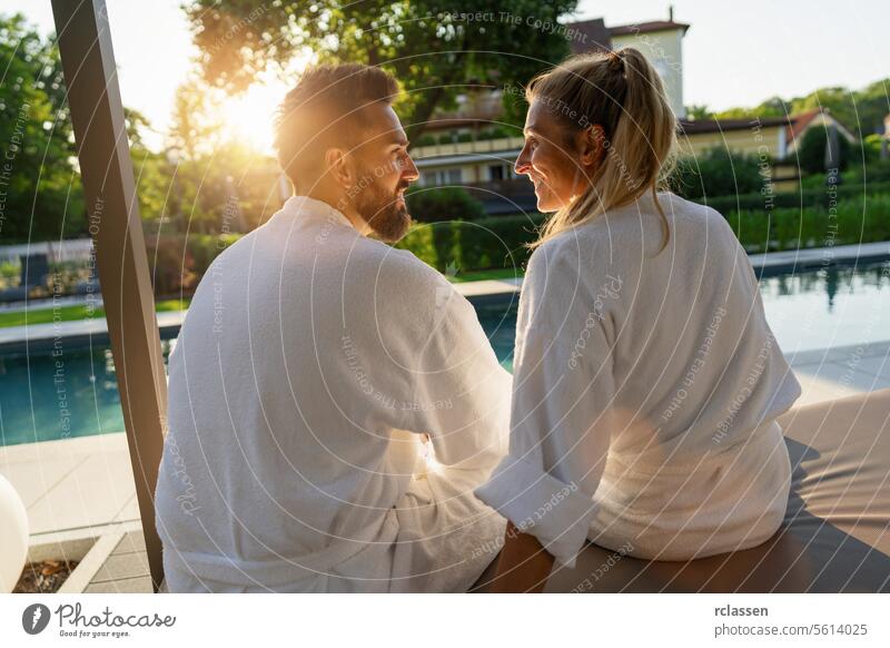 Verliebtes Paar in weißen Bademänteln genießt den Sonnenuntergang an einem Pool in einem Wellness-Hotel Resort Liebe weiße Gewänder Erholung Freizeit Genuss Spa