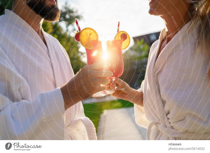 Paar in weißen Bademänteln stößt mit roten Cocktails an, während die Sonne im Spa-Hotel untergeht Röstung rote Cocktails weiße Gewänder Sonnenlicht im Freien