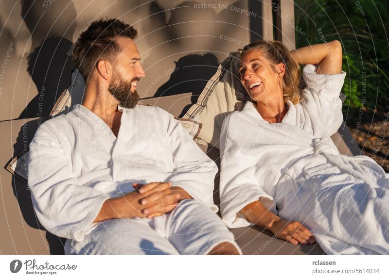 Paar in weißen Bademänteln, das sich auf einer Liege im Sonnenlicht in einem Wellness-Hotel ausruht und lacht Bankliege sich[Akk] entspannen Resort Liebe