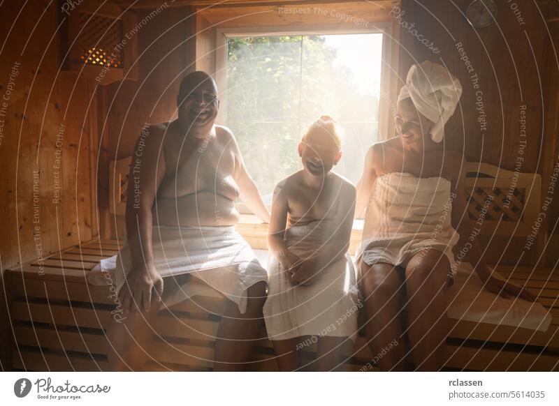 Familie in einer finnischen Sauna lacht zusammen, Mann links, Frau mit Handtuch auf dem Kopf rechts, Kind in der Mitte im Wellnesshotel Finnland Spa-Wellness