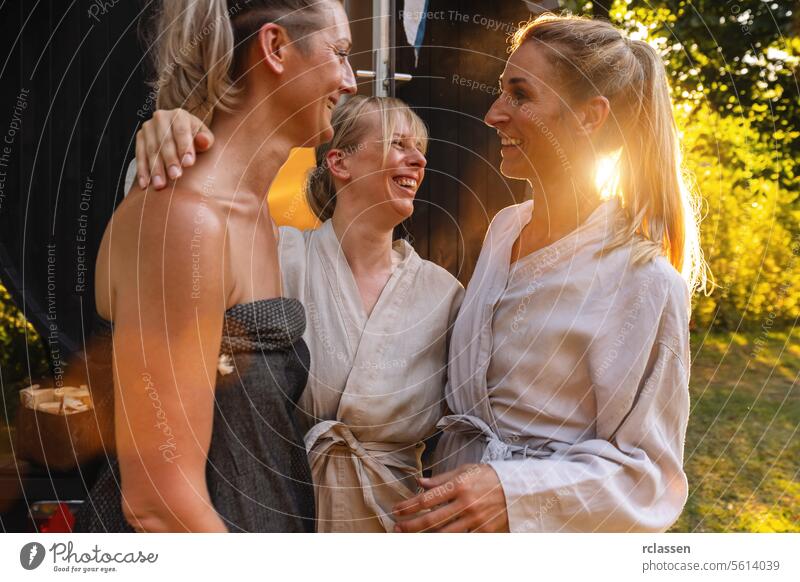 Drei fröhliche Menschen in Bademänteln vor einem finnischen Saunafass, lachend und plaudernd Mobile glückliche Menschen Roben finnische Sauna Lachen Erholung