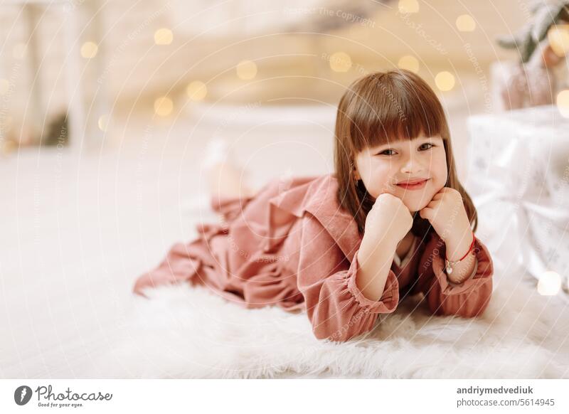 kleine niedliche Kind Mädchen in rosa Kleid auf weißen pelzigen Teppich in dekoriert Weihnachten Zimmer Interieur und Blick in die Kamera liegen. Schöne Bokeh. Glückliche Familie Neujahr, Xmas Konzept