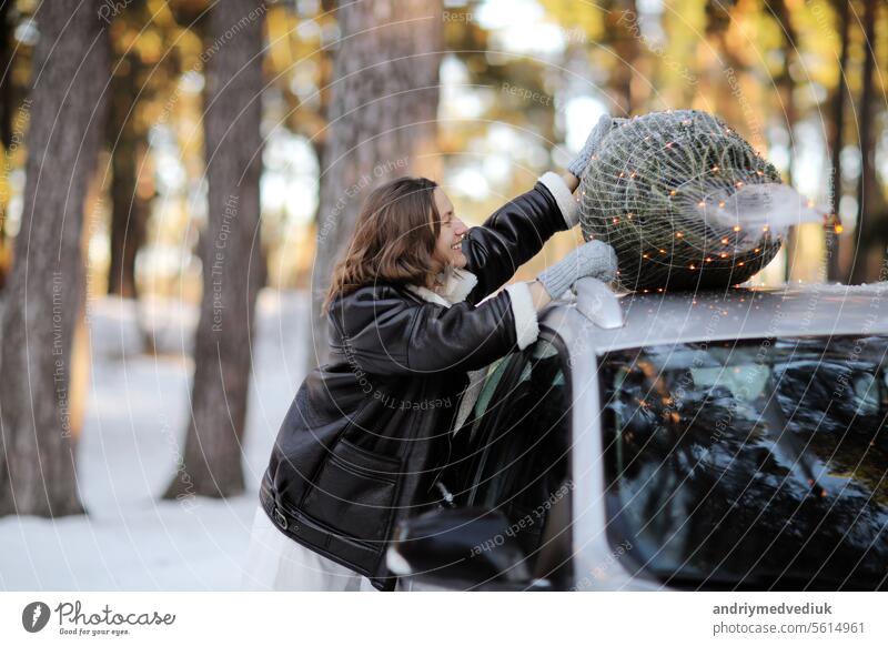 Lächelnde kaukasische Frau in Wintermantel und Fäustlinge in der Nähe von Auto mit beleuchtetem Weihnachtsbaum auf einem Dach in der Natur in verschneiten Wald. Konzept des Feierns von Neujahrsferien. Idee von Xmas Stimmung und Spaß