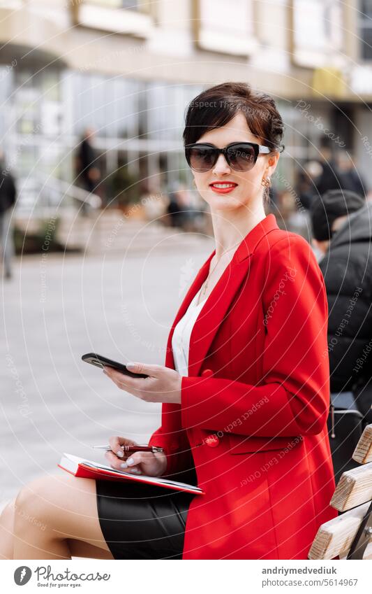 Anwalt erfolgreiche Geschäftsfrau in rotem Blazer und Sonnenbrille sitzt auf Bank in der Stadt Straße sprechen auf Smartphone. Lächelnde Frau macht Business-Anruf auf dem Handy und macht Notizen in Notebook im Freien.