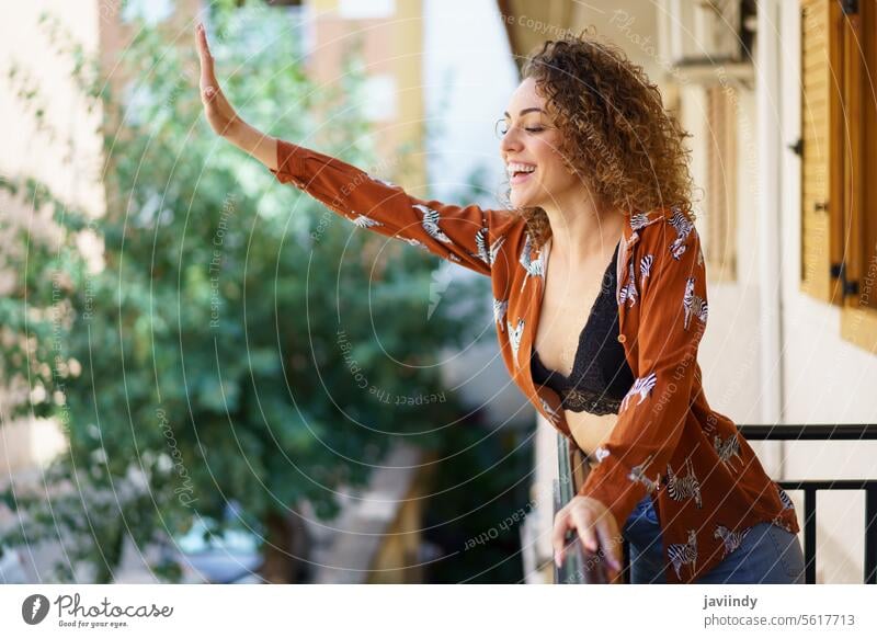 Fröhliche Frau zeigt einen Gruß, während sie auf dem Balkon ihres Hauses steht gestikulieren Lächeln zeigen Individualität heiter Outfit krause Haare Optimist
