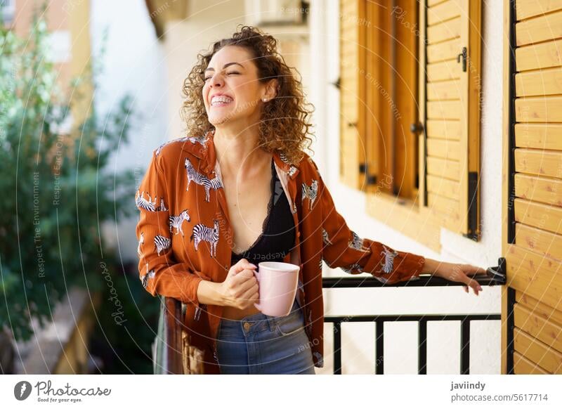 Fröhliche junge Frau hält eine Kaffeetasse in der Hand und lehnt sich auf dem Balkon eines Glück Stil Lächeln trendy heiter Tasse Terrasse brünett genießen