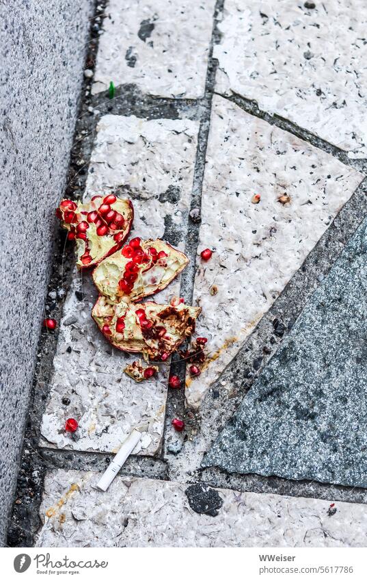 Ein kaputter Granatapfel liegt neben einer Kippe auf der Straße im Dreck Müll Überfluss Asphalt dreckig fallen matschig Nahrung Luxus achtlos Nahrungsmittel