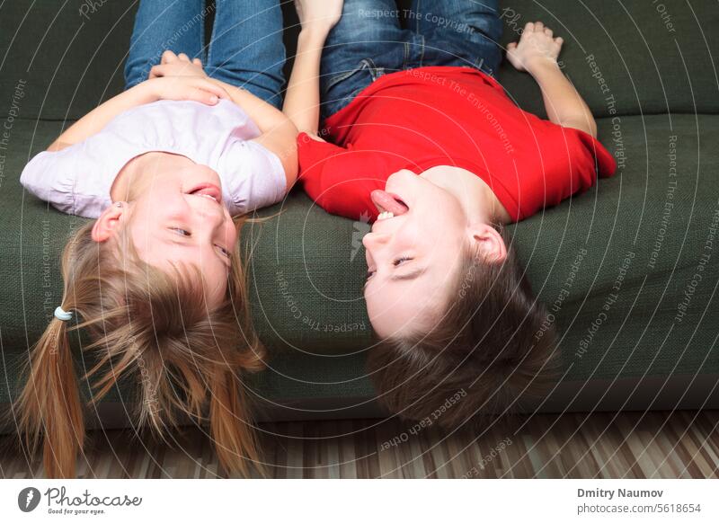 Bruder und Schwester in Freizeitkleidung liegen auf einem grünen Sofa zu Hause und strecken ihre Zungen aus, um sich gegenseitig zu necken Halbgeschwister Junge