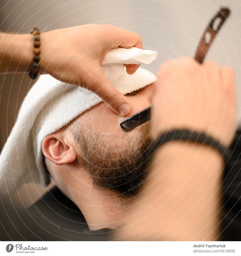 Ein Barbier rasiert die Wange eines bärtigen Kunden mit einem gefährlichen Rasiermesser. Rasieren der Kontur des Bartes für die richtige Form. Rasur Vollbart