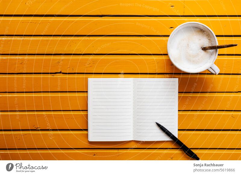 Eine Tasse Kaffee und ein Notizbuch auf einem gelben Holztisch. Draufsicht Kugelschreiber Tisch Getränk Frühstück heiß Morgen Koffein Kaffeepause Tagebuch