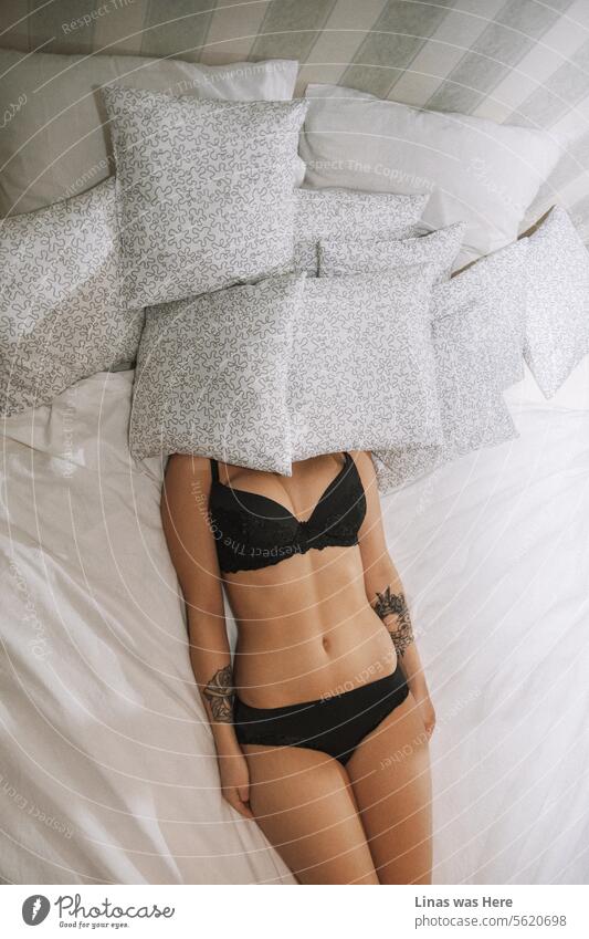 Ein wunderschönes Dessous-Modell in schwarzem Slip und schwarzem BH verdeckt ihr Gesicht unter den Kissen. Eine schlafende, eingefärbte Schönheit fühlt sich entspannt in ihrer eigenen Haut und in ihrer eigenen Unterwäsche. Ihr sonnengeküsster Körper liegt anmutig auf dem Bett.