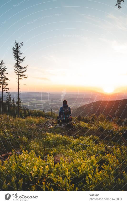 Abenteurer, der auf einem Baumstumpf auf dem Gipfel eines Berges sitzt und einen orange-roten Sonnenuntergang beobachtet. Genießt den Erfolg. Beskiden, Tschechische Republik
