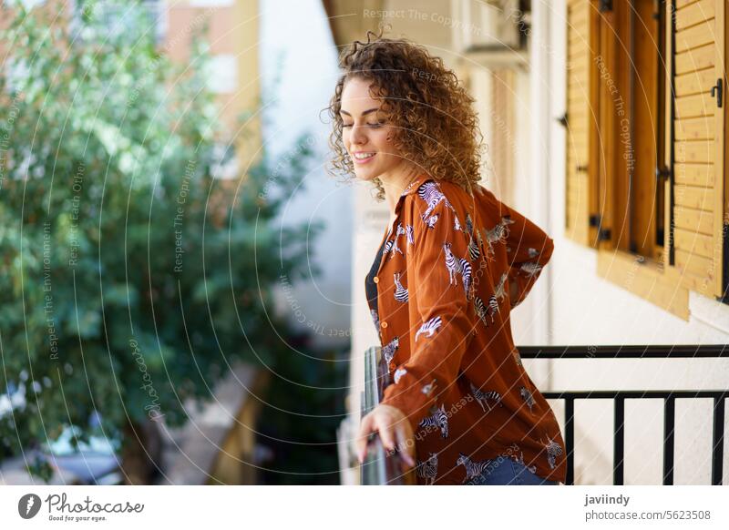 Glückliche junge Frau in Freizeitkleidung mit lockigem Haar steht auf einem Balkon Lächeln positiv lässig heiter charmant Optimist froh Vorschein angenehm