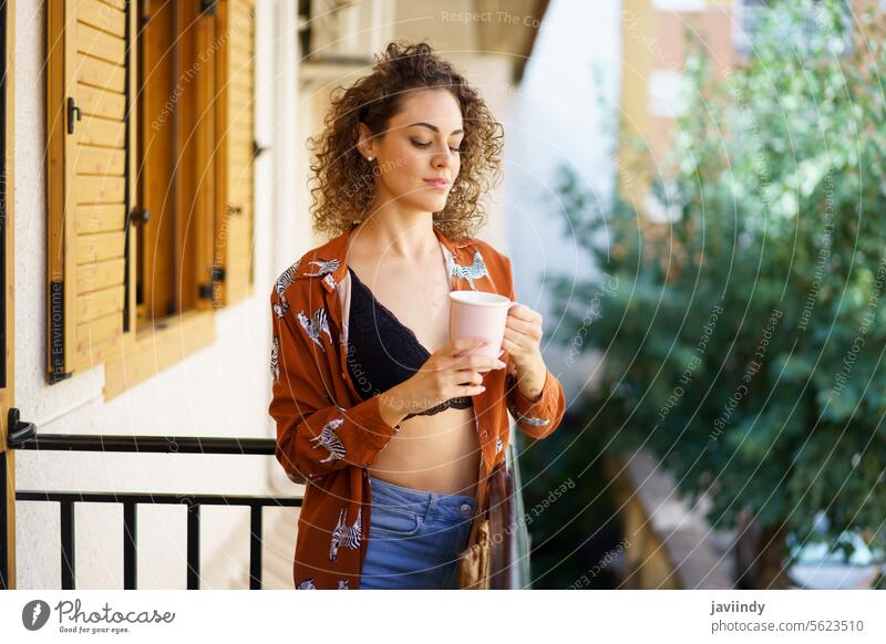 Junge stilvolle lächelnde Frau mit einer Tasse Kaffee auf dem Balkon zu Hause stehend Lächeln Mode trinken Outfit krause Haare genießen selbstbewusst Inhalt
