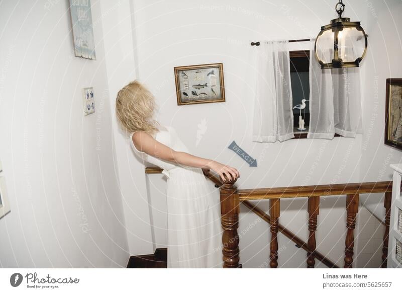 Eine atemberaubende blonde Frau in einem eleganten weißen Kleid geht eine Holztreppe hinunter, die sich in einem charmanten Haus am Meer befindet. Ein schönes Mädchen mit lockigem blondem Haar posiert mit Anmut und verleiht der Szene einen Hauch von Einzigartigkeit, da sie ein Hochzeitskleid trägt.