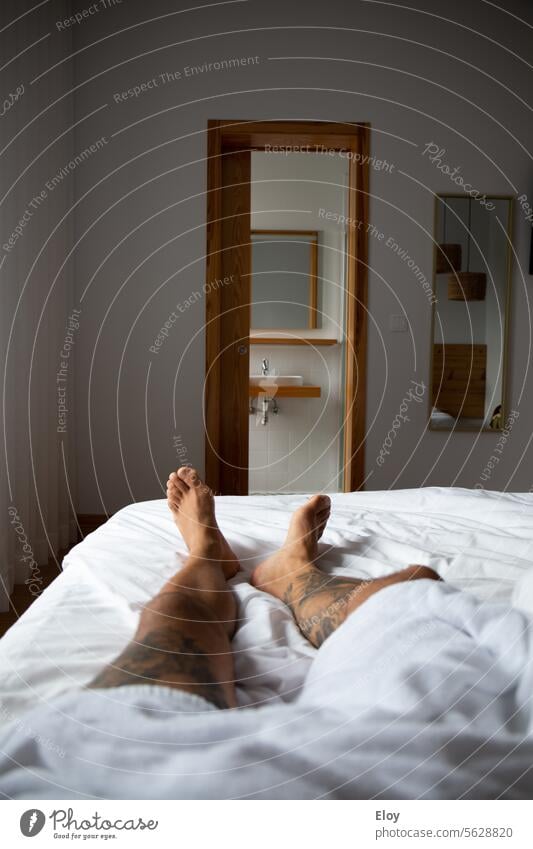 Mann ruht sich in einem Hotelzimmer aus, Nahaufnahme der Beine eines tätowierten Mannes, der mit einem weißen Handtuch auf dem Bett liegt, im Hintergrund sind ein Badezimmer und ein Spiegel zu sehen