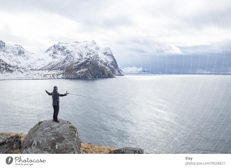 Mann in Winterlandschaft, behüteter Mann mit offenen Armen auf einem Felsen vor dem dunklen Meer stehend, im Hintergrund sieht man die schneebedeckten Berge, es ist kalt und der Himmel ist bewölkt