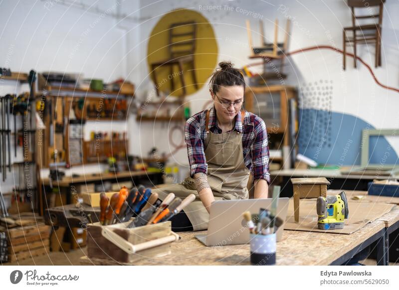 Junge Handwerkerin mit Laptop in ihrer Werkstatt Möbel Zimmerer wiederherstellend Schreinerei Holz Stuhl Gebäude Inszenierung Renovierung reparierend Nutzholz