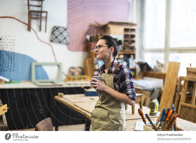 Porträt einer jungen Schreinerin mit einer Tasse Kaffee in der Hand in ihrer Werkstatt Möbel Zimmerer wiederherstellend Schreinerei Holz Stuhl Gebäude