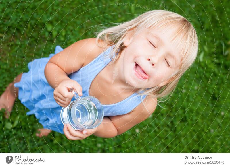 Hohe Winkel Ansicht Schuss von blonden kleinen Mädchen trägt blaues Kleid hält Glas Wasser lecken ihre Lippen mit ihren Augen in einem Sommergarten geschlossen