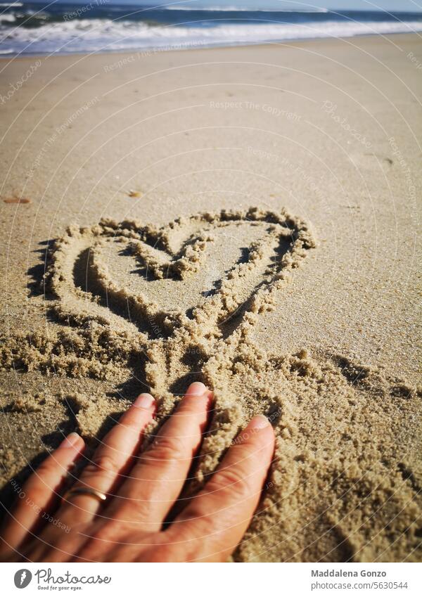 Eine Hand zeichnet gerade ein Herz in den Sand am Strand Zeichnung Fingerzeichnung Markerstellung Meer Ehering Liebe Heirat winken Hochzeit romantisch Romantik
