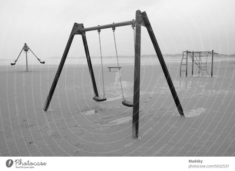 Verlassene Spielgeräte mit Bank bei Regen und Sturm am Strandspielplatz am Strand von St. Peter-Ording im Kreis Nordfriesland in Schleswig-Holstein im Herbst an der Nordseeküste in Schwarzweiß