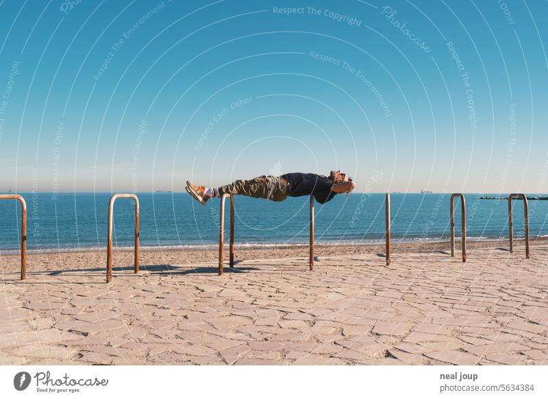 Mann liegt als Planke bequem über zwei Fahrradbügel und optisch auf dem Meereshorizont Paradoxon Widerspruch Entspannung Yoga starr stabil Spannung Kraft