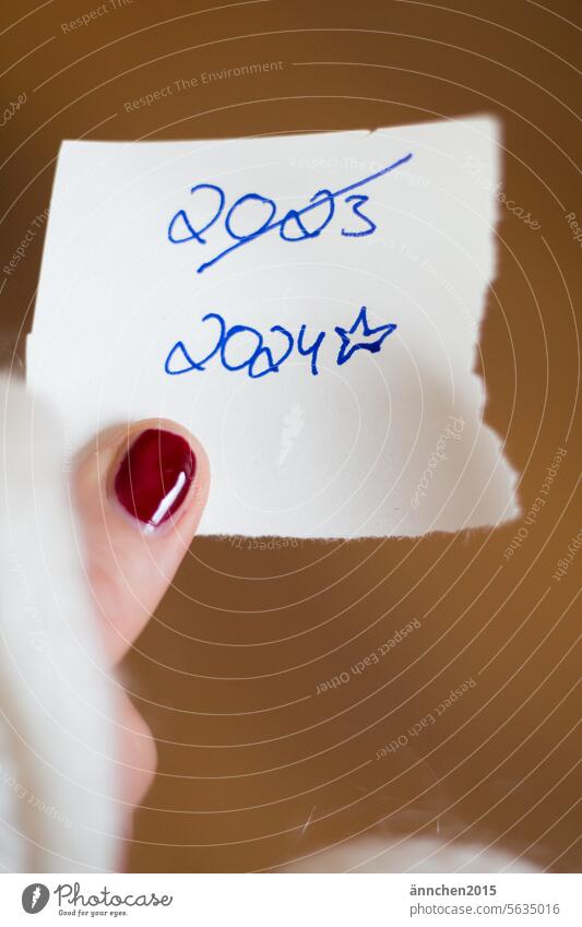 ein Zettel mit den Daten 2023 und 2024 wird von einem Finger mit rot lackiertem Nagel gehalten Silvester Neujahr Fingernagel Nagellack neues Jahr feiern