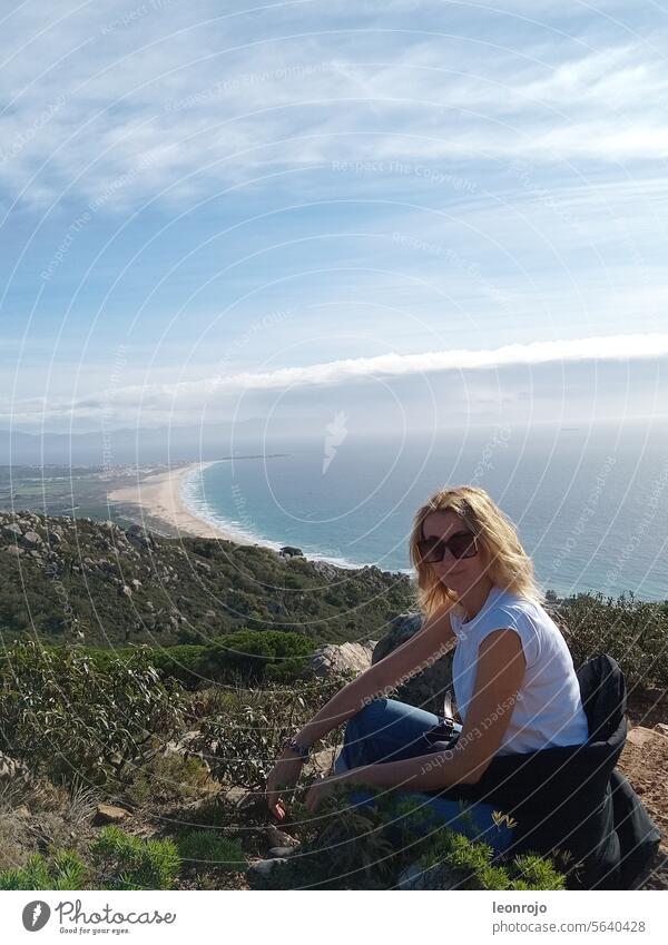 Eine schöne Frau mit Tshirt und Sonnenbrille sitzt tagsüber auf einem Berg und genießt die Ruhe und das Panorama. Im Hintergrund sieht man einen langen Strand und den Ozean.