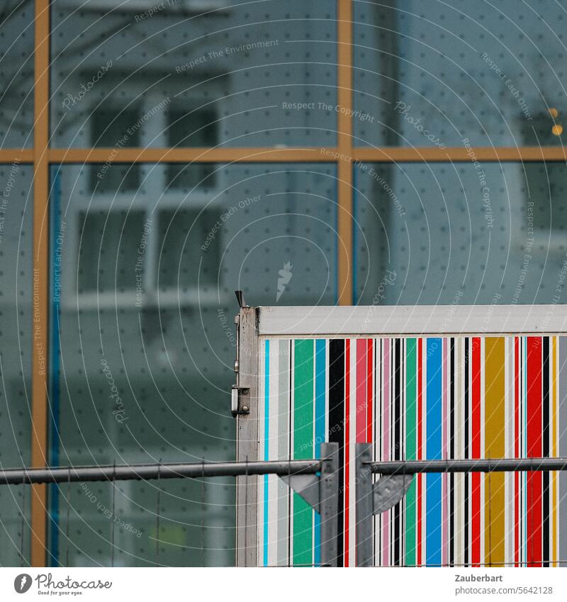 Ein Container mit bunten Barcode-Streifen steht vor einer Glasfassade mit geometrischen Formen, Spiegelung Fassade Stadt Baustelle Gegensatz graphisch abstrakt