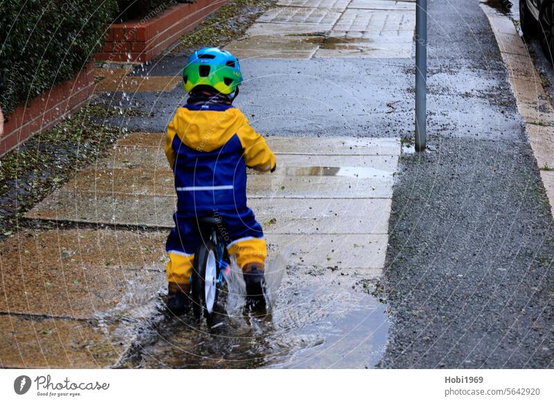 Kleines Kind fährt mit einem Laufrad durch eine Pfütze Rad laufen Fahrrad Fahrradfahren Wasser spritzen Regen Spaß Regenjacke Jacke Helm Fahrradhelm Gehweg