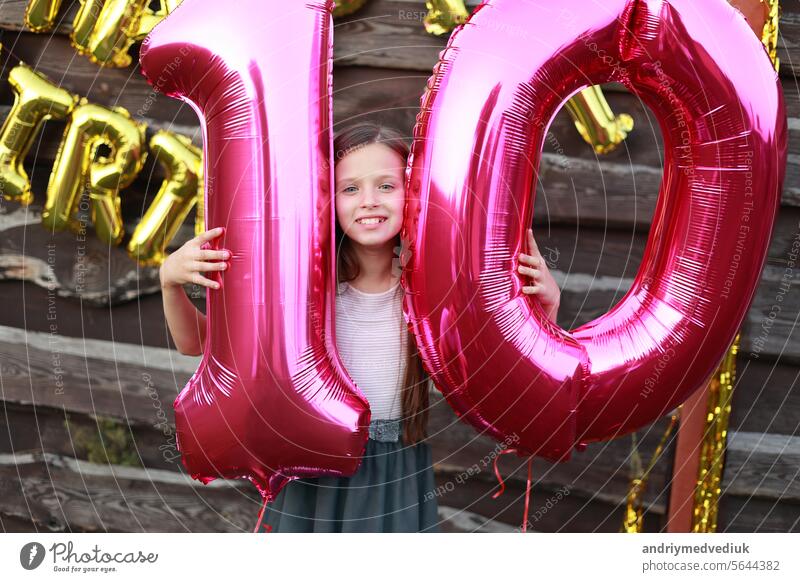 Fröhlich kleines Mädchen hält zwei große rosa Folienballons, Nummer 10 im Freien auf hölzernen Wand Hintergrund mit goldenen Ballons Aufschrift alles Gute zum Geburtstag. Zehn Jahre Jahrestag Feier Partei zu Hause