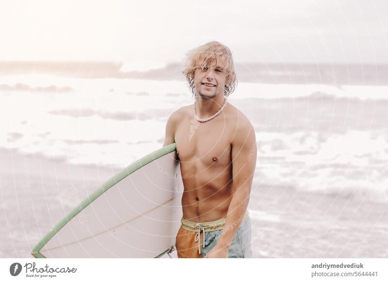 Handsome fit jungen blonden Mann mit Mock-up-Surfbrett wartet auf Welle zum Surfen vor Ort am Meer Ozean Strand mit schwarzem Sand und schaut in die Kamera. Konzept der Sport, Fitness, Freiheit, Glück, neue moderne Leben