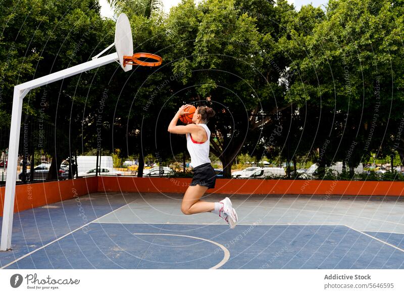 Seitenansicht in voller Länge einer entschlossenen jungen Frau in Sportkleidung, die springt und den Ball wirft, um einen Korb zu machen, während sie auf dem Platz spielt