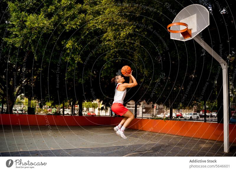 Seitenansicht in voller Länge einer entschlossenen jungen Frau in Sportkleidung, die springt und den Ball wirft, um einen Korb zu machen, während sie auf dem Platz spielt