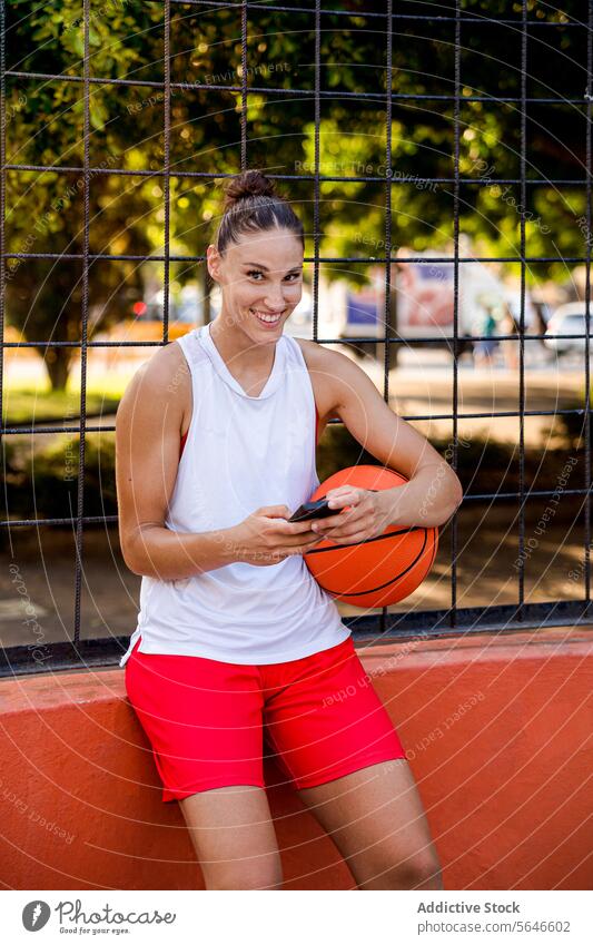 Lächelnde junge Basketballspielerin, die einen Ball hält und ein Smartphone benutzt, während sie in die Kamera auf einem Spielplatz an einem sonnigen Tag schaut