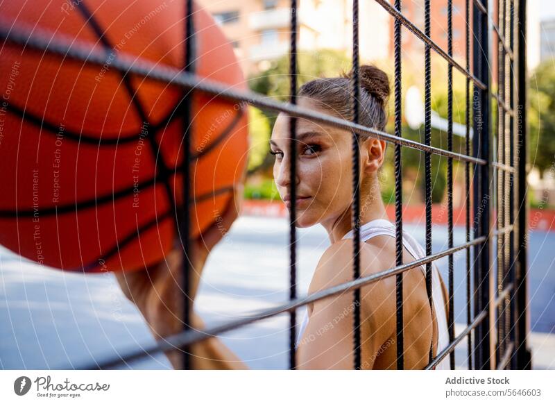 Selbstbewusste junge Sportlerin in Sportkleidung, die einen Basketball hält und in die Kamera schaut, gesehen durch den Zaun eines Spielplatzes Ball Athlet