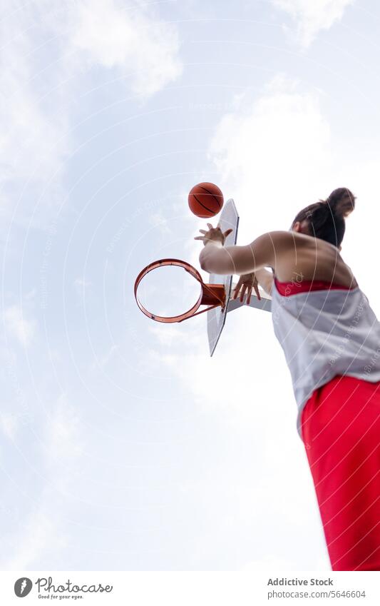 Ausschnitt von unten einer nicht erkennbaren weiblichen Basketballspielerin in Sportkleidung, die springt und den Ball in einen Korb vor einem bewölkten Himmel wirft