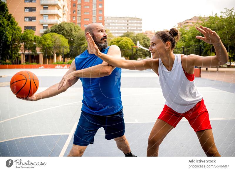 Entschlossener Mann und Frau in Sportkleidung spielen Basketball auf einem Spielplatz in einer Stadt mit Gebäuden und Bäumen im Hintergrund Sportler Ball wehren