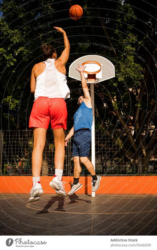 Rückenansicht einer Sportlerin in Sportkleidung, die einen Basketball in Richtung Korb schießt, und eines Mannes, der versucht, sich auf dem Platz gegen den Himmel zu verteidigen