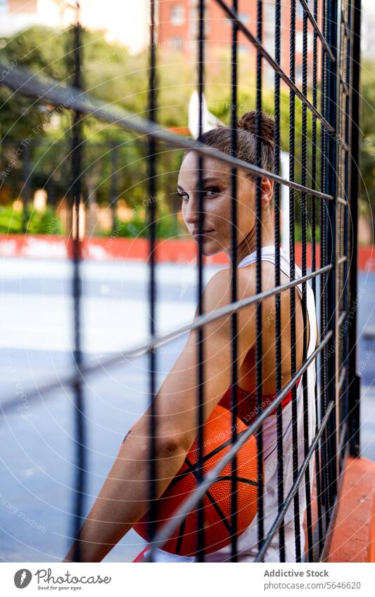 Selbstbewusste junge Sportlerin in Sportkleidung, die einen Basketball hält und in die Kamera schaut, gesehen durch den Zaun eines Spielplatzes Ball Athlet