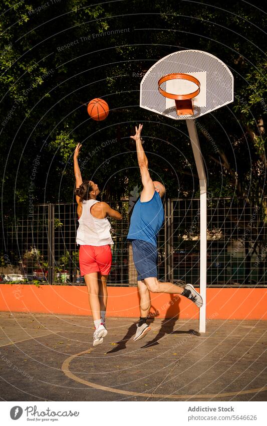 Rückenansicht einer Sportlerin in Sportkleidung, die einen Basketball in Richtung Korb schießt, und eines Mannes, der versucht, sich auf dem Platz gegen den Himmel zu verteidigen