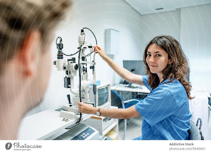 Glückliche junge Ärztin, die in der Nähe eines Spaltlampen-Biomikroskops mit einem anonymen Mann im Krankenhaus steht Frau Arzt Lächeln Uniform Stethoskop
