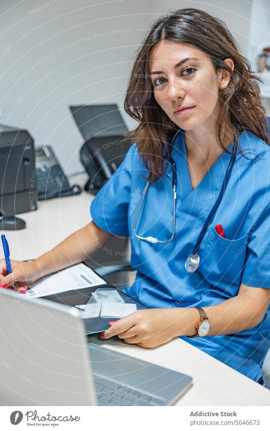 Junge Ärztin sitzt am Tisch mit Laptop und Smartphone und macht Notizen auf dem Klemmbrett Frau Arzt schreiben Lächeln Zwischenablage Stethoskop Schreibstift