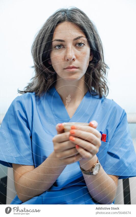 Junge Ärztin hält eine Tasse Kaffee und schaut in die Kamera, während sie sitzt Frau Arzt Uniform Krankenhaus medizinisch Gesundheitswesen Spezialist sitzen