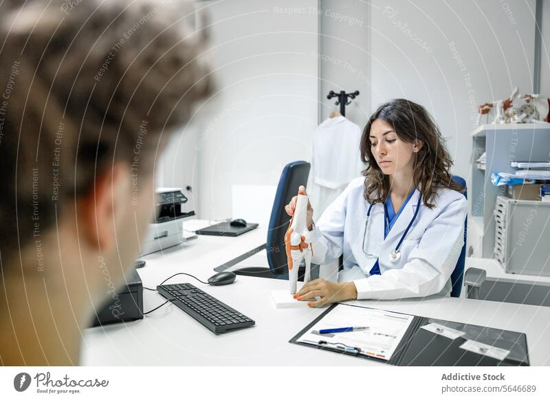 Eine Ärztin sitzt am Tisch mit einer Sehnen- und Muskelfigur in einer Klinik und zeigt sie einem nicht erkennbaren männlichen Patienten Frau Arzt Uniform Mann