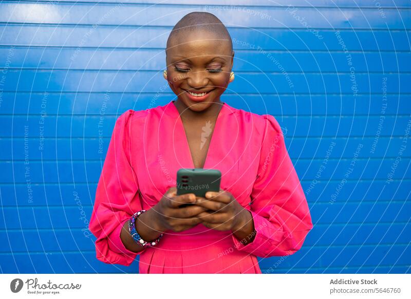 Fröhliche schwarze Frau mit rasiertem Kopf in rosafarbener Kleidung, die ein Smartphone vor einer blauen Wand benutzt benutzend positiv Browsen Lächeln Glück